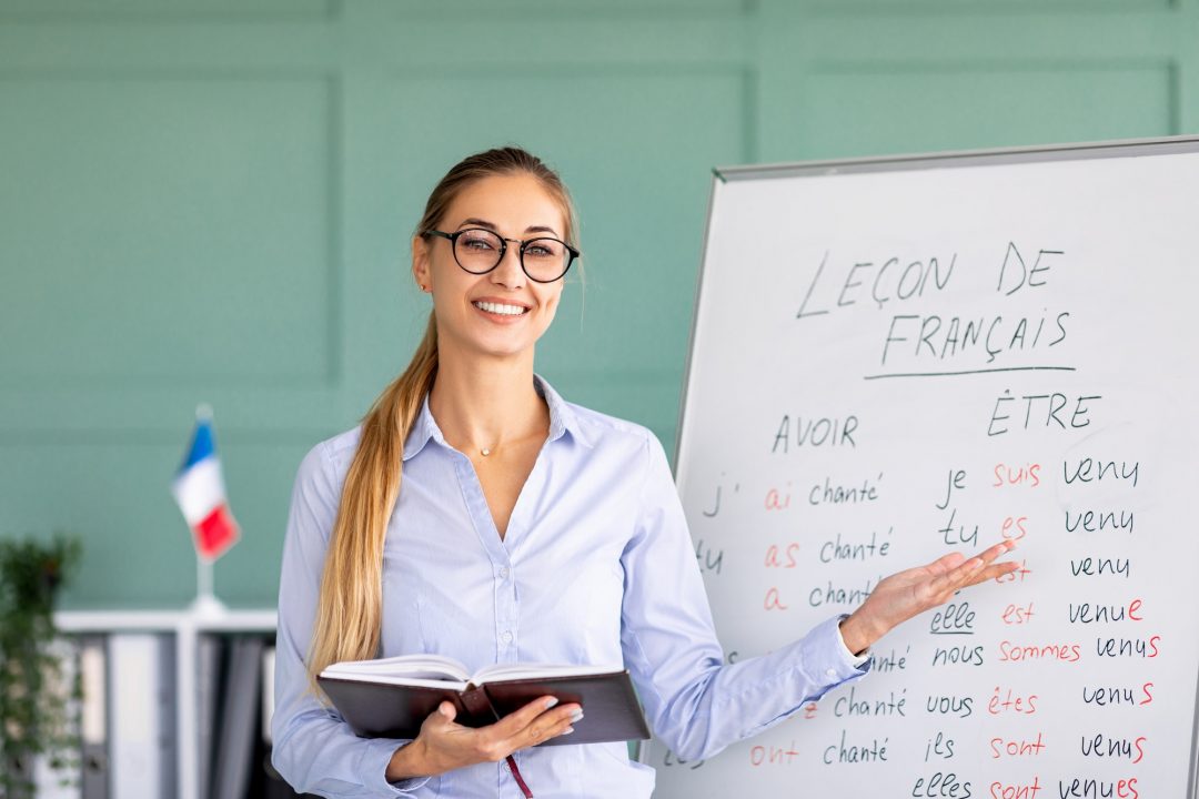 happy french teacher explaining foreign language rules near blackboard indoors and smiling at camera